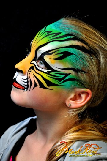 girl in painted on mask over the top of her face of a tigers face in yellow green and blue